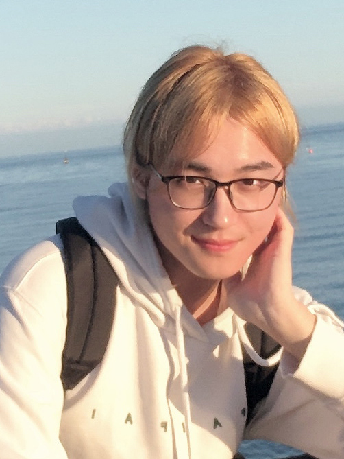 A person with short blonde hair wearing glasses and a white hoodie, sitting outdoors with a calm ocean in the background.